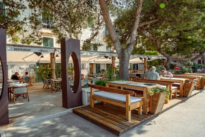 Drveni stolovi među zelenim krošnjama ispred ulaza u restoran